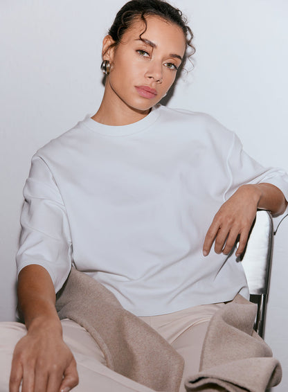 A woman with curly hair pulled back, wearing a white Luxe Boxy Short Sleeve top, seated and looking at the camera [M-170cm]