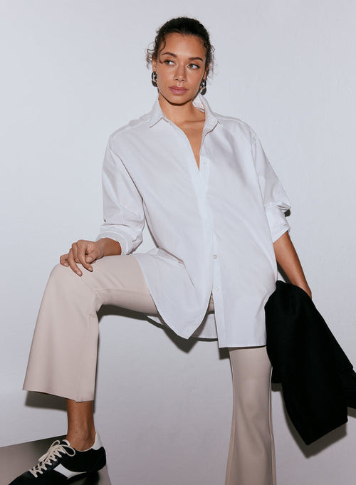 A woman wears a white oxford shirt, light beige trousers, and black sneakers, holding a black jacket, posing with one leg raised on a surface [M-170cm]