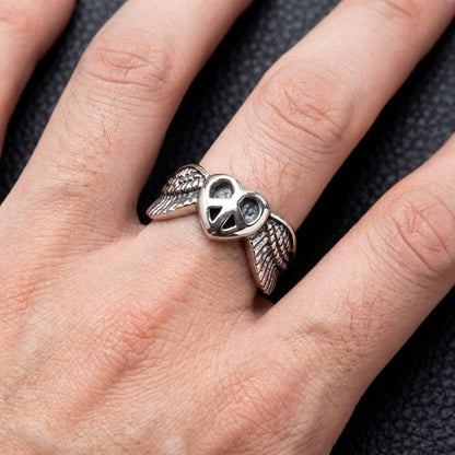 Silver ring with a heart peace sign and wings worn on a hand against dark leather.